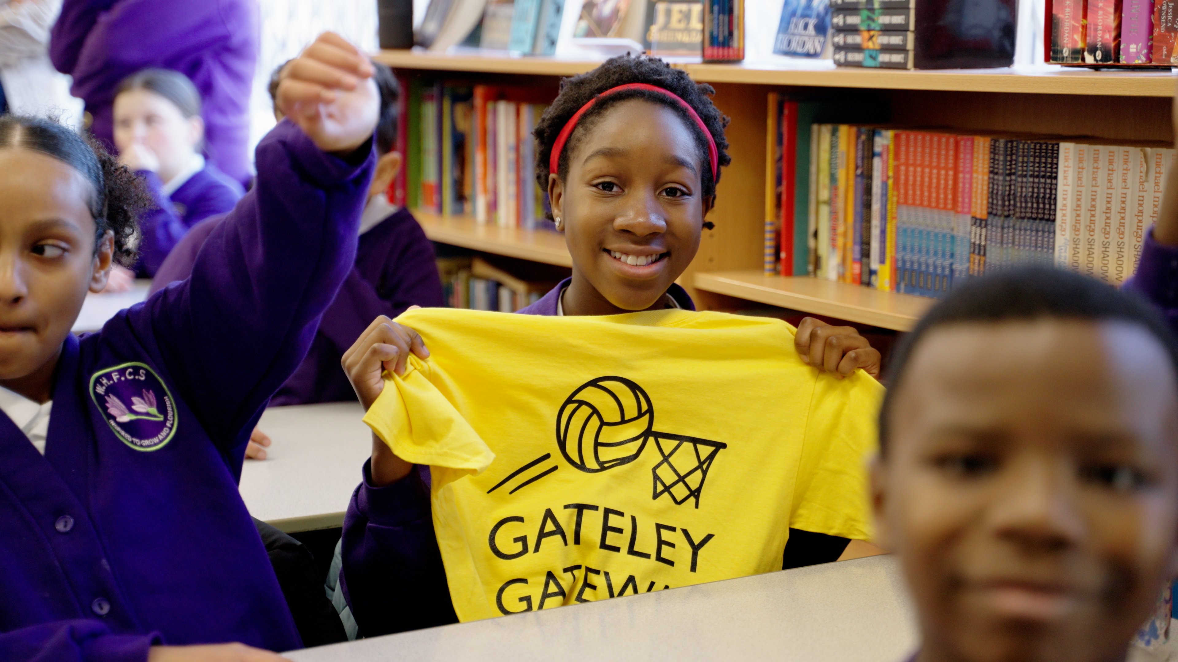 One of the children smiles to camera holding up a Gateley Gateway to Netball t-shirt